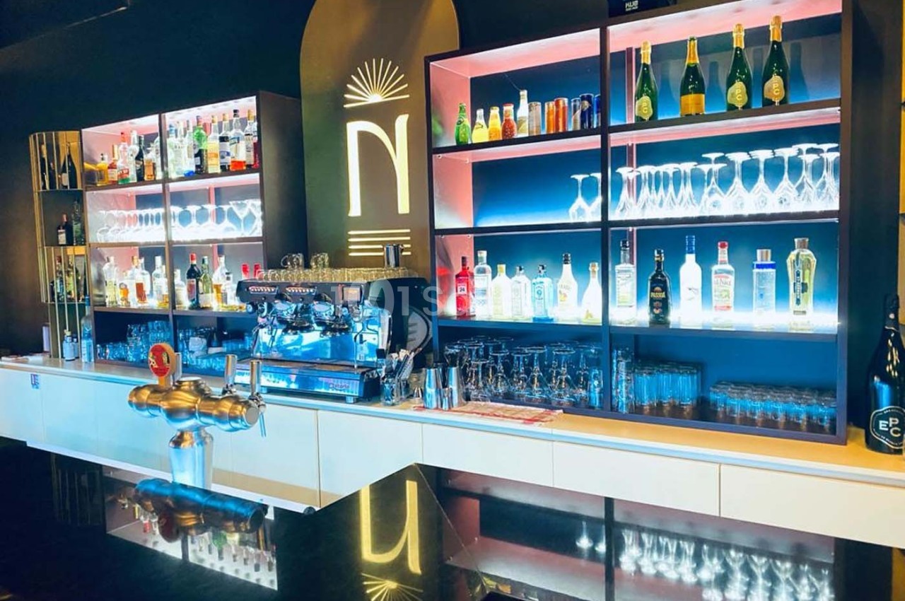 Bar moderne avec étagères éclairées présentant des bouteilles d'alcool, verres et machine à café en arrière-plan.