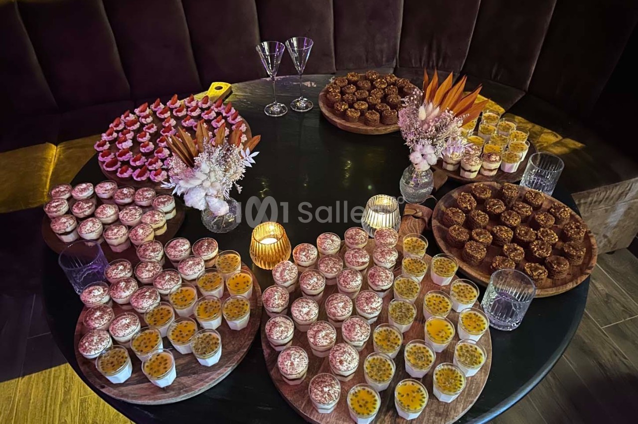 Table ronde garnie de desserts variés, incluant verrines, pâtisseries et décorations florales, éclairée par des bougies.