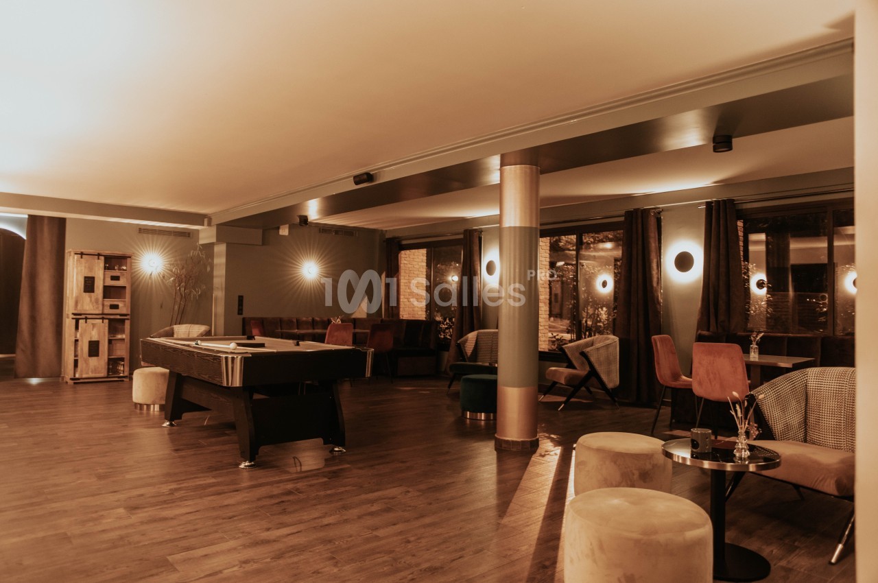 Salle intérieure avec parquet, billard, tables, chaises et éclairage tamisé, ambiance chaleureuse et moderne.