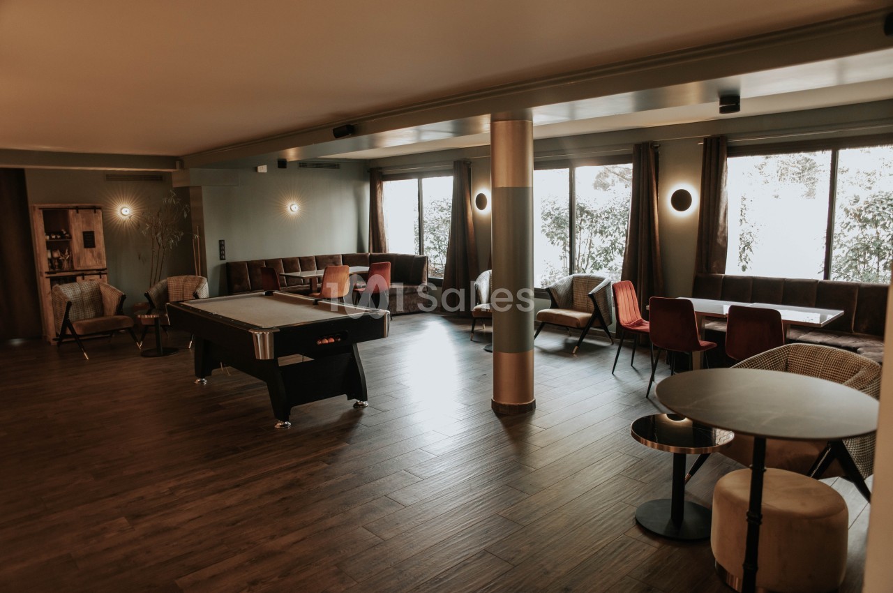 Salle lumineuse avec parquet, billard, tables et chaises, décorée de tons neutres et grandes fenêtres donnant sur l…