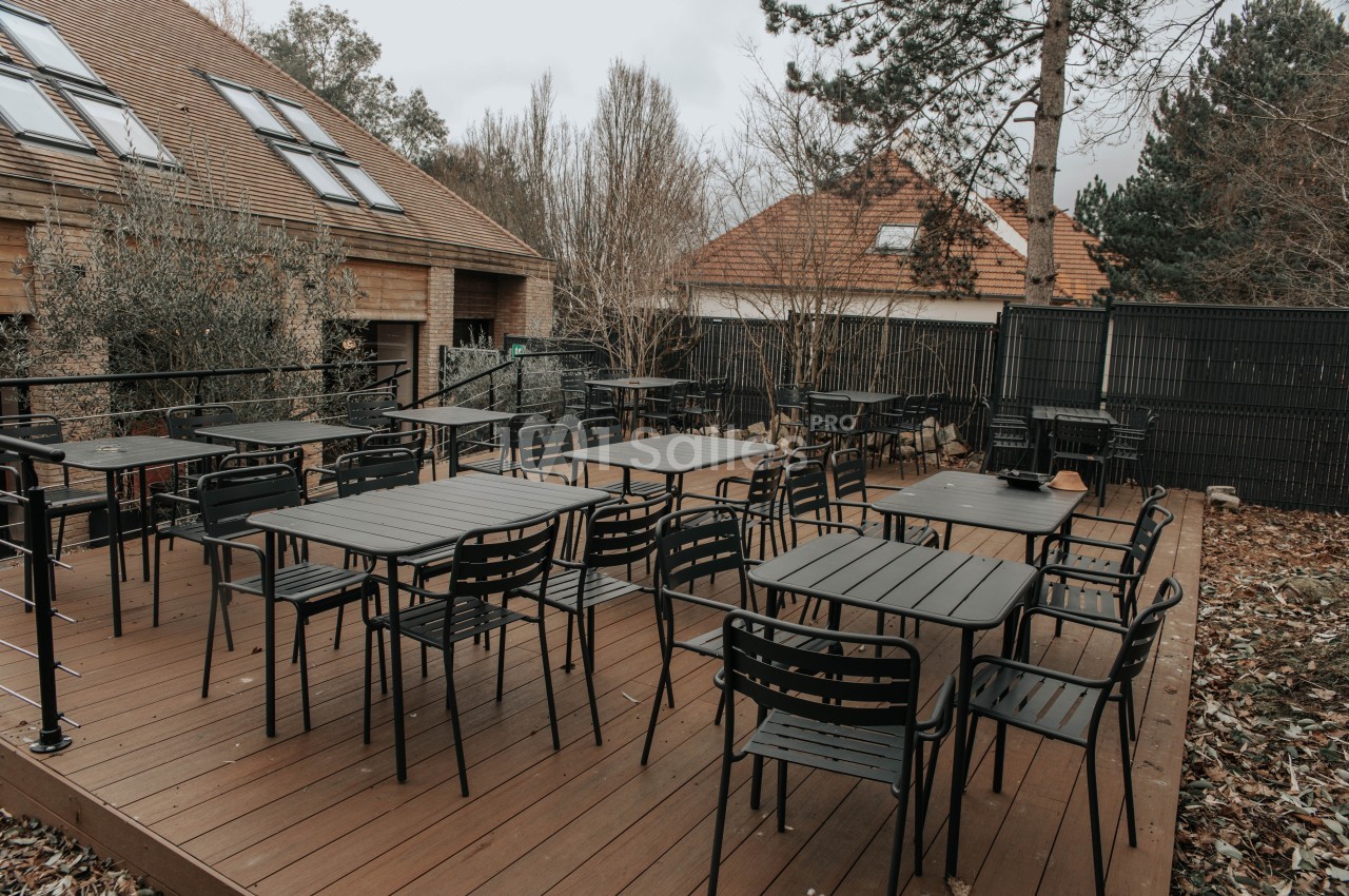 Terrasse en bois avec des tables et chaises noires, entourée de végétation et de bâtiments résidentiels.