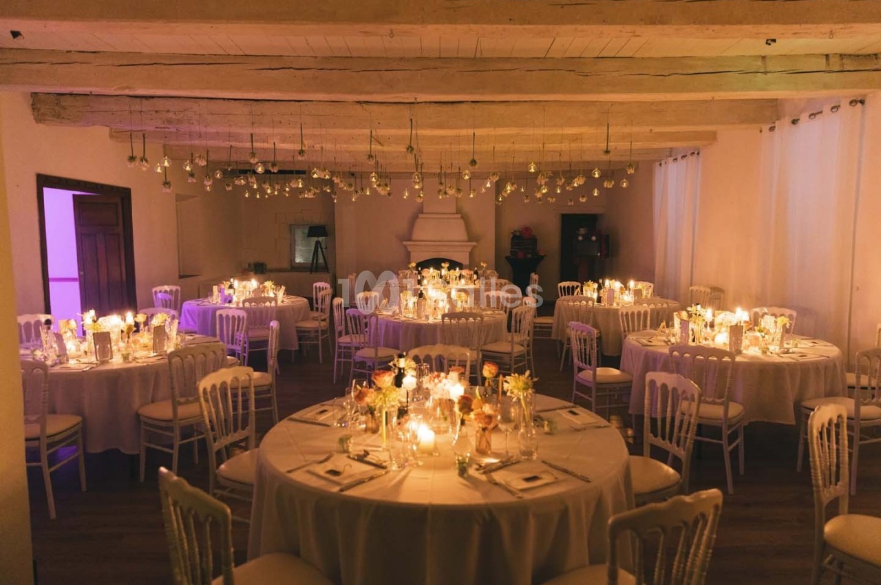 Salle de réception décorée avec des tables rondes, nappes blanches, bougies et guirlandes lumineuses suspendues.