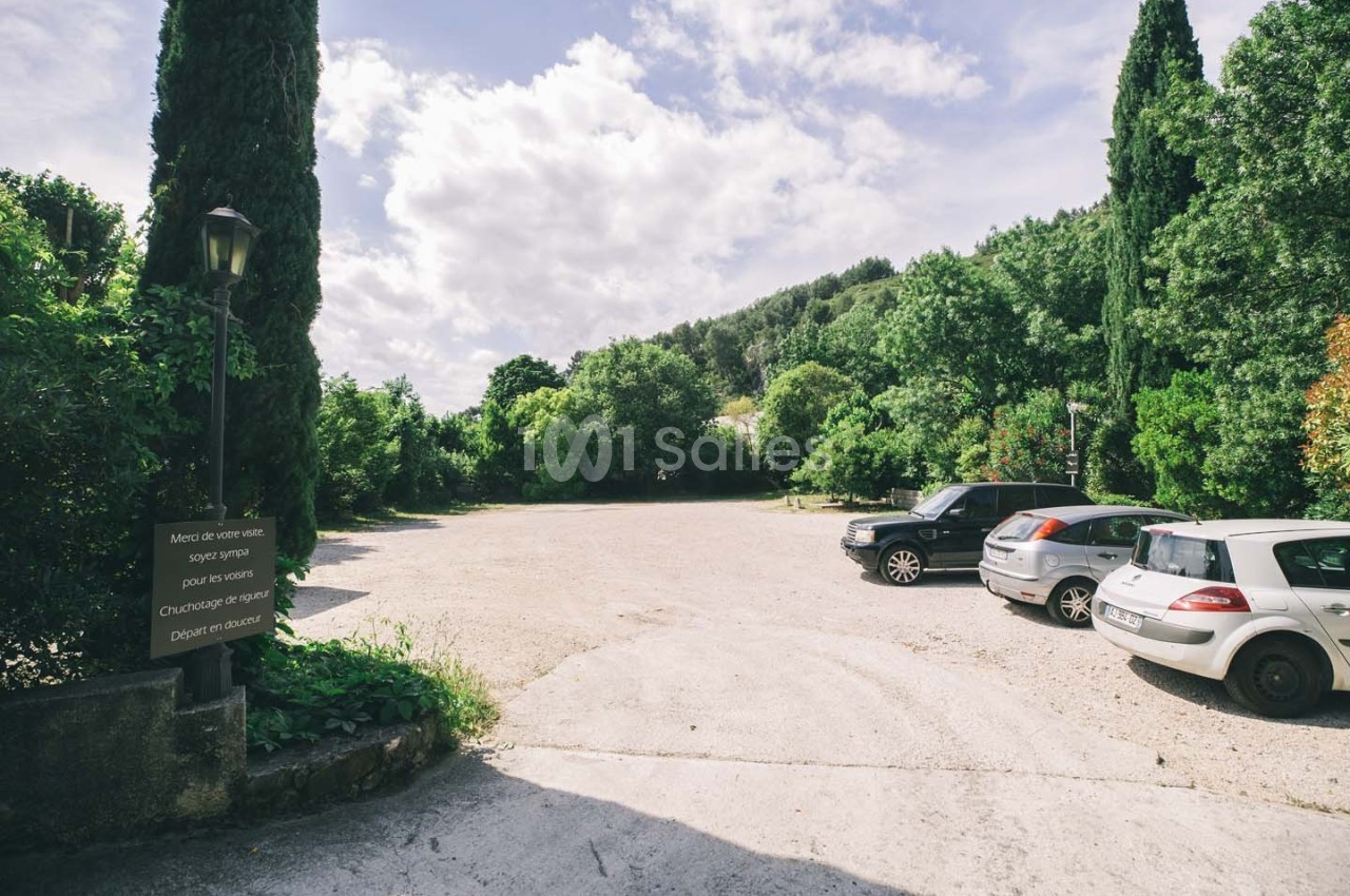 Parking extérieur entouré de végétation, avec quelques voitures stationnées et un panneau à gauche.