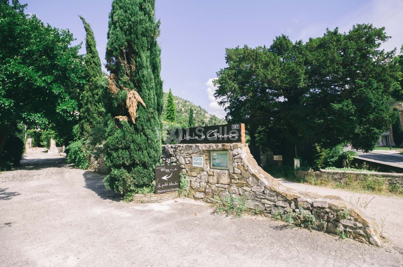 Entrée en pierre d'un domaine avec des panneaux indiquant ’Le Hameau des Baux’, entourée de végétation méditerranéenne.