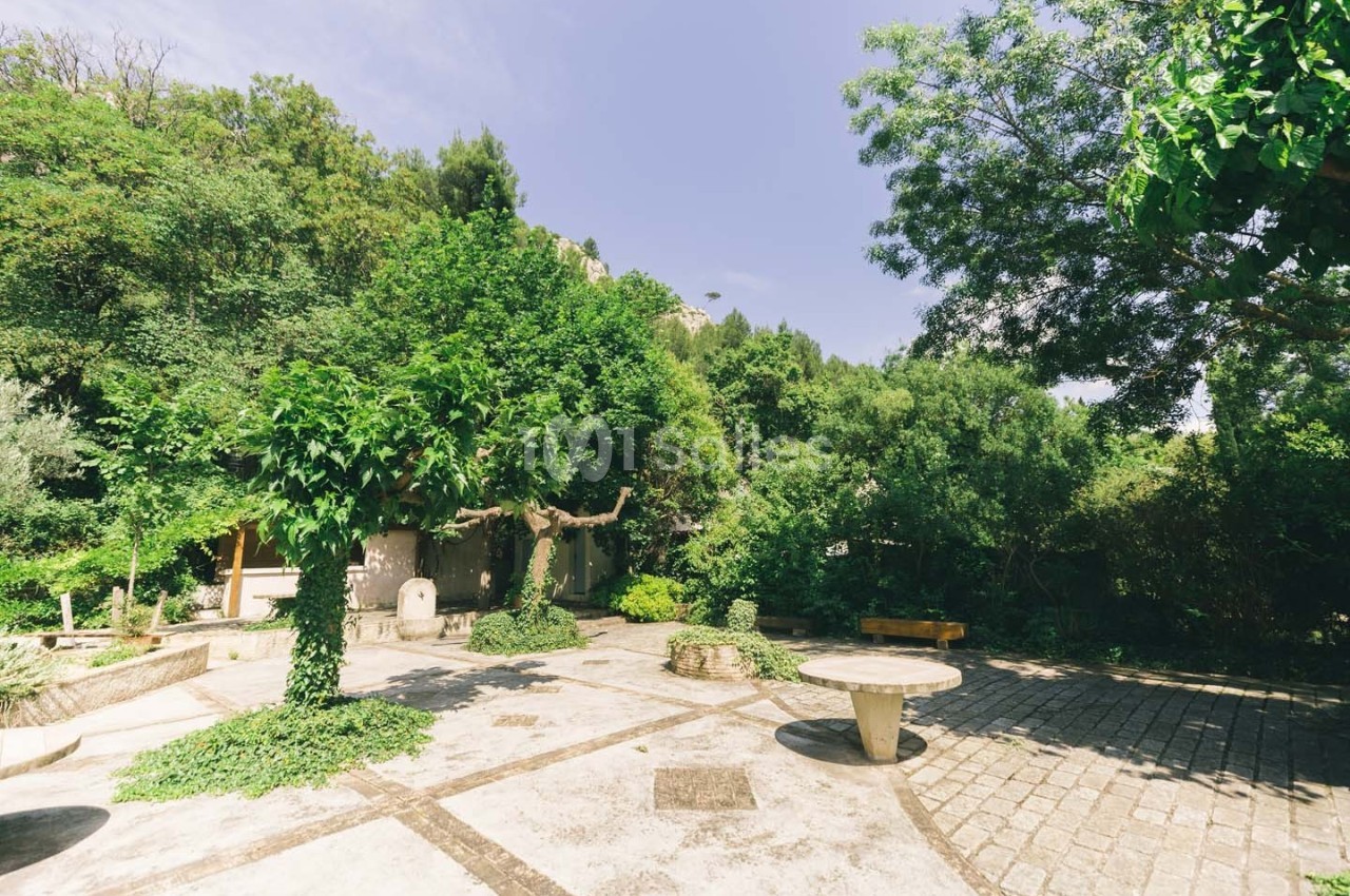 Cour extérieure ensoleillée entourée de végétation dense, avec des arbres, des bancs et une table en pierre.