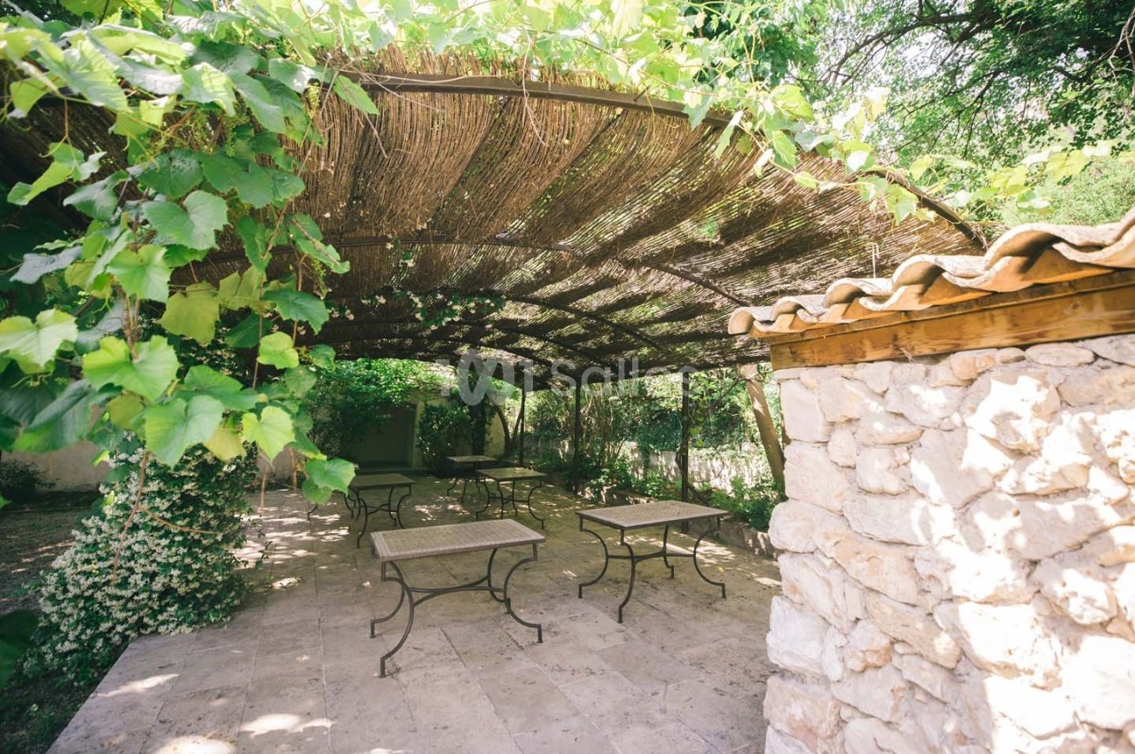 Terrasse ombragée avec pergola en bois et feuillage, tables en métal et mur en pierre dans un cadre naturel.