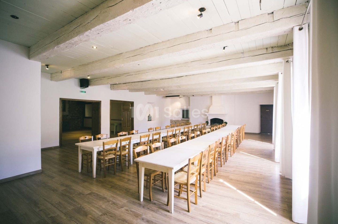 Salle lumineuse avec de longues tables en bois, des chaises assorties et un plafond avec poutres apparentes.