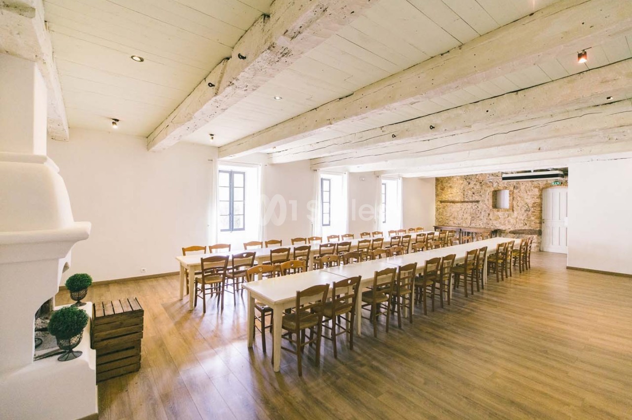 Salle lumineuse avec poutres apparentes, grandes fenêtres, murs en pierre et tables alignées entourées de chaises en bois.