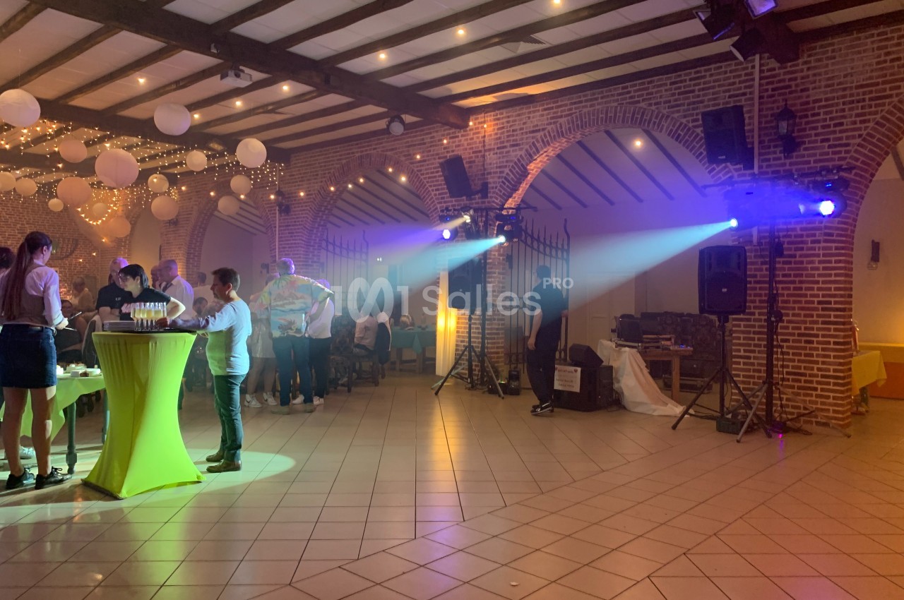 Salle de réception éclairée avec des guirlandes lumineuses, des invités debout près de tables hautes et une ambiance festive.