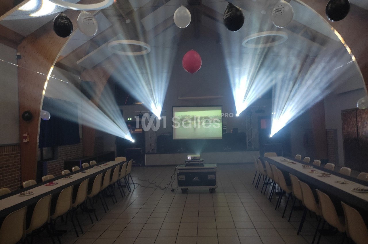 Salle décorée avec des ballons, tables alignées et projecteur diffusant une image, éclairée par des faisceaux lumineux.