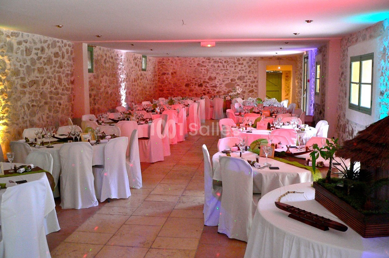 Salle de réception en pierre avec tables rondes dressées, nappes blanches et éclairage coloré.