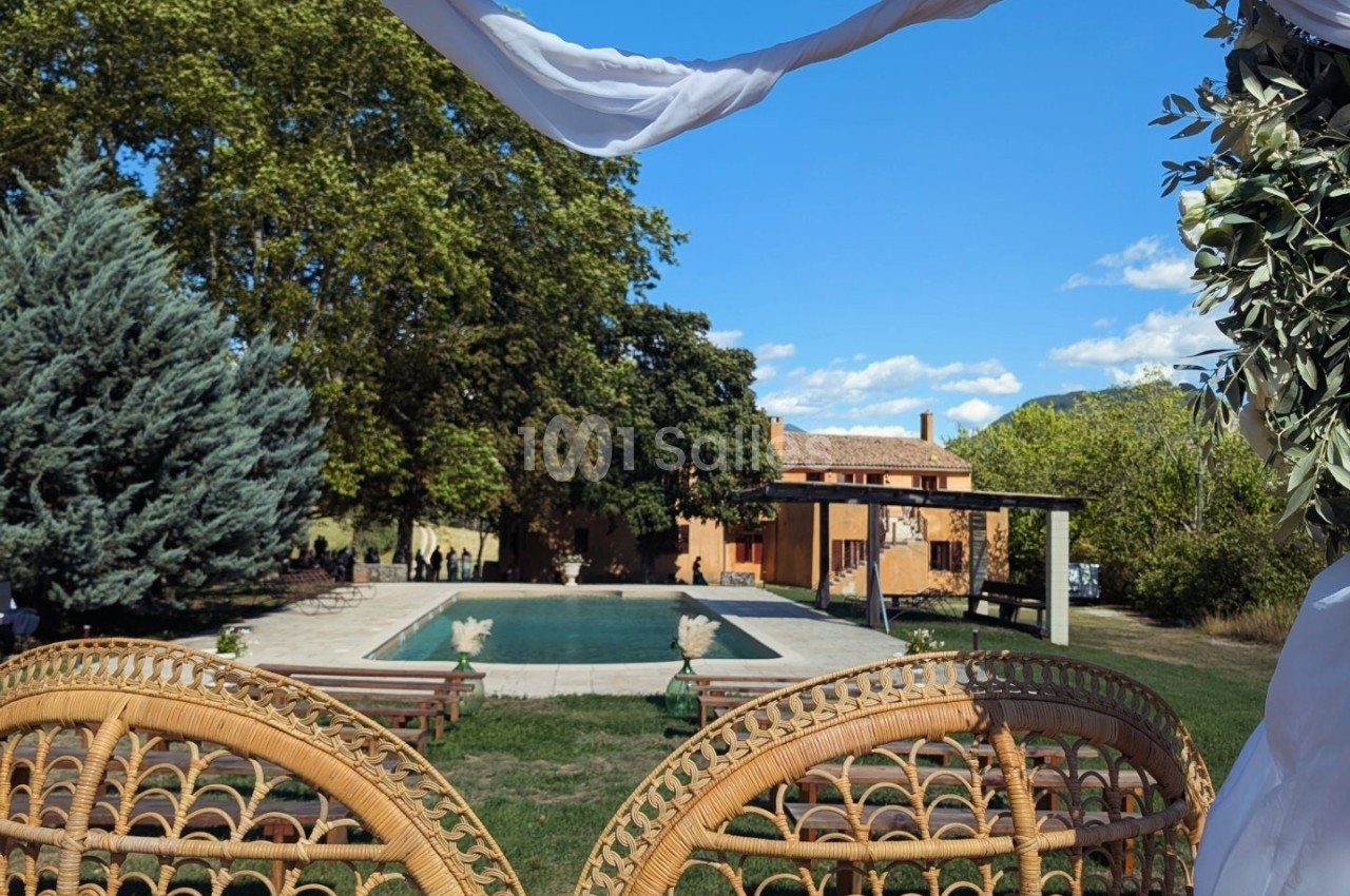 Vue sur une piscine entourée de verdure et de chaises en rotin, avec une maison en arrière-plan sous un ciel bleu.