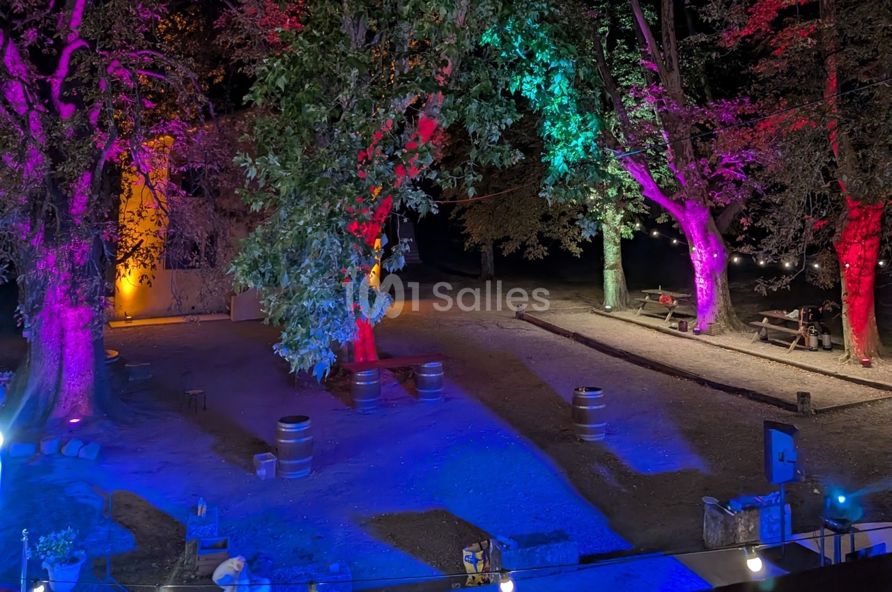 Éclairage coloré illuminant des arbres et un espace extérieur avec des tonneaux et des tables en soirée.