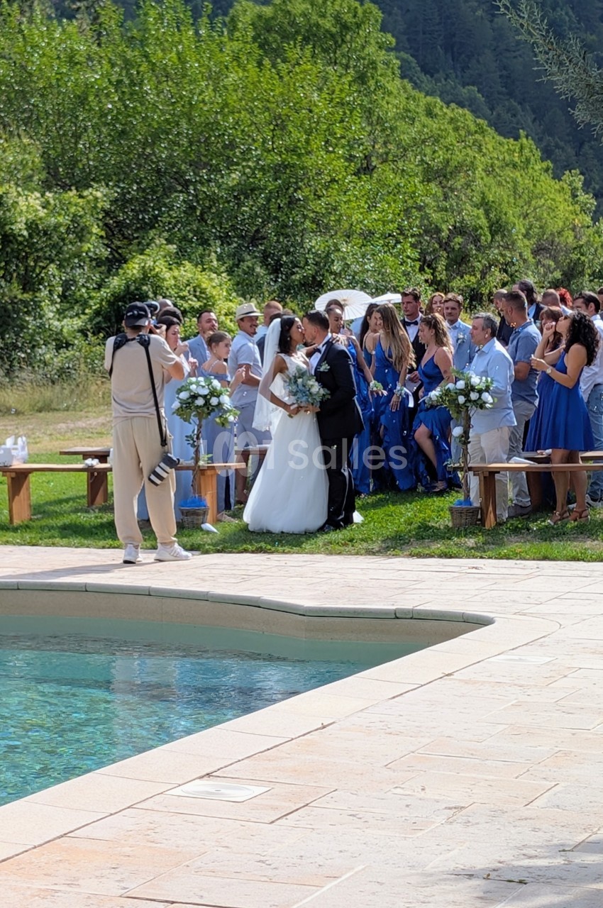 Un couple de mariés pose devant des invités en tenue élégante près d'une piscine, dans un cadre naturel.
