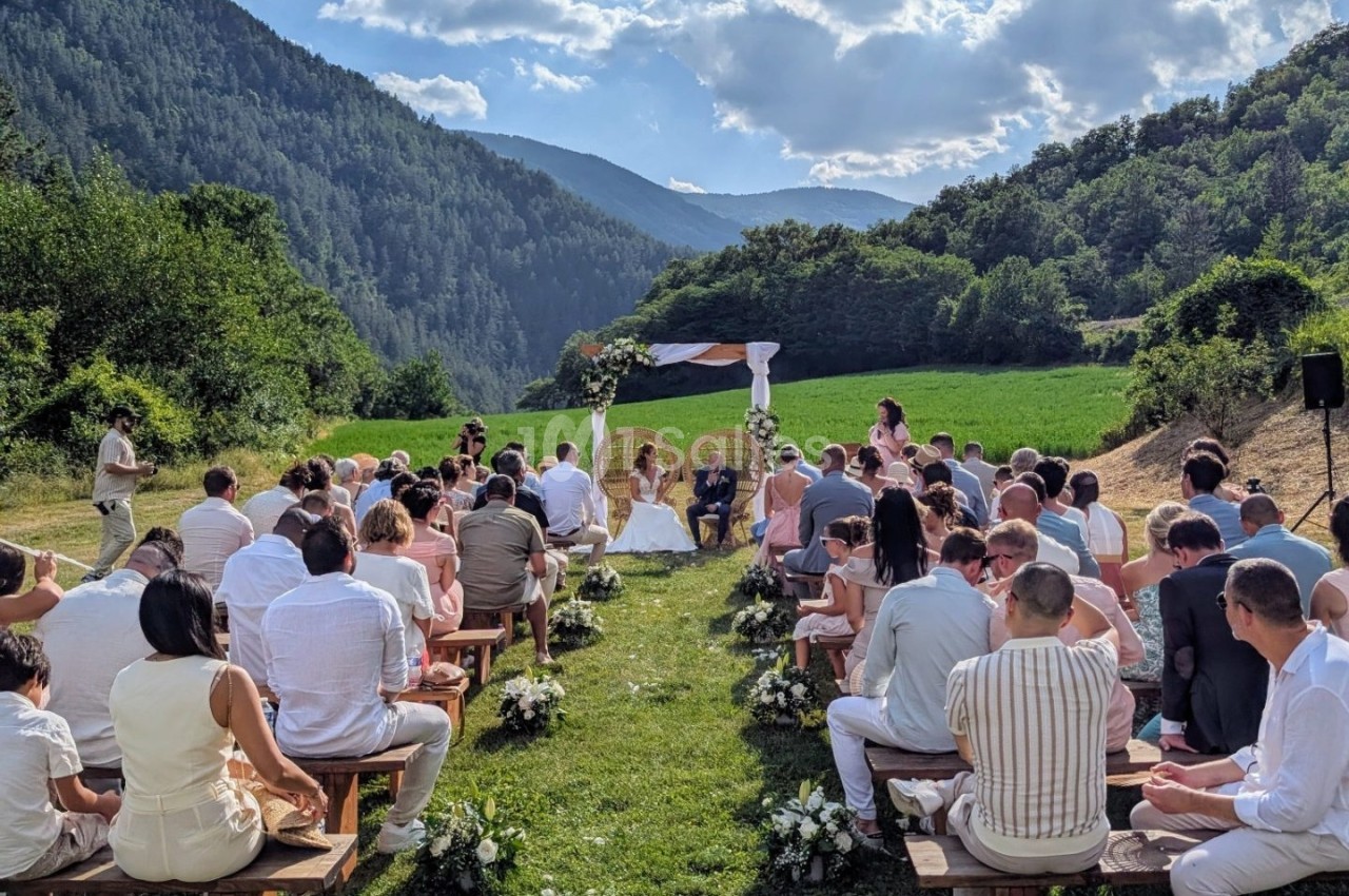 Cérémonie de mariage en plein air dans un cadre montagneux, avec des invités assis et un couple sous une arche fleurie.