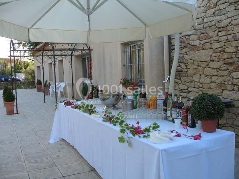 Table dressée en extérieur avec boissons, verres et décoration florale, sous un grand parasol près d'un mur en pierre.