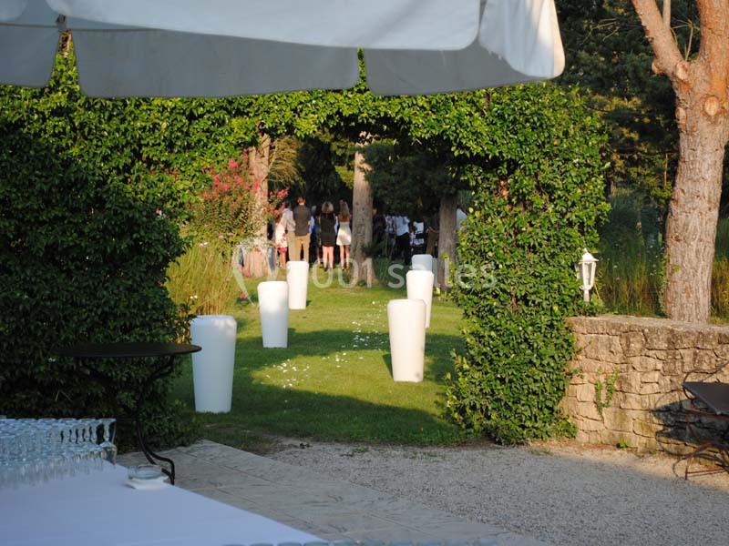 Vue d'un jardin avec une arche végétale, des invités rassemblés au loin et des décorations blanches sur la pelouse.