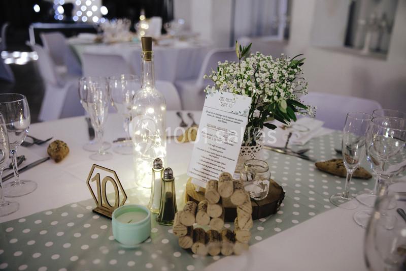 Table décorée pour un événement avec une bouteille lumineuse, un menu, des fleurs blanches et des accessoires en liège.