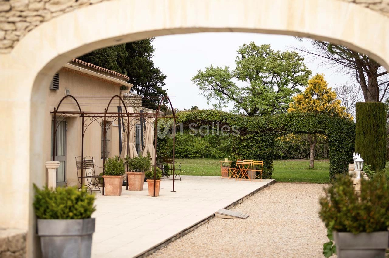 Terrasse avec pergolas en métal, pots de plantes, et vue sur un jardin arboré avec une table en bois jaune.