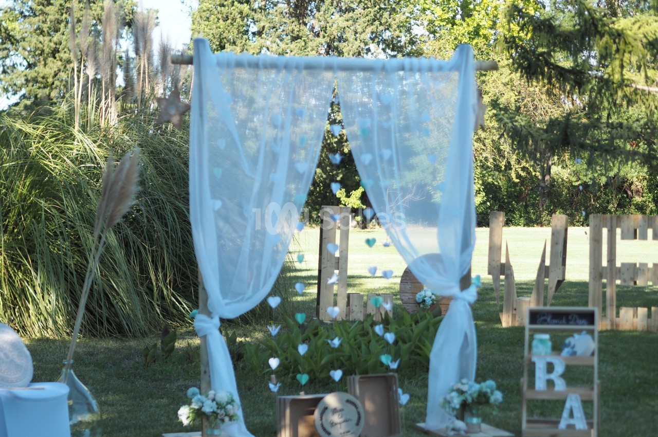 Arche de mariage décorée de voilages blancs, fleurs et éléments en bois, installée dans un jardin verdoyant.