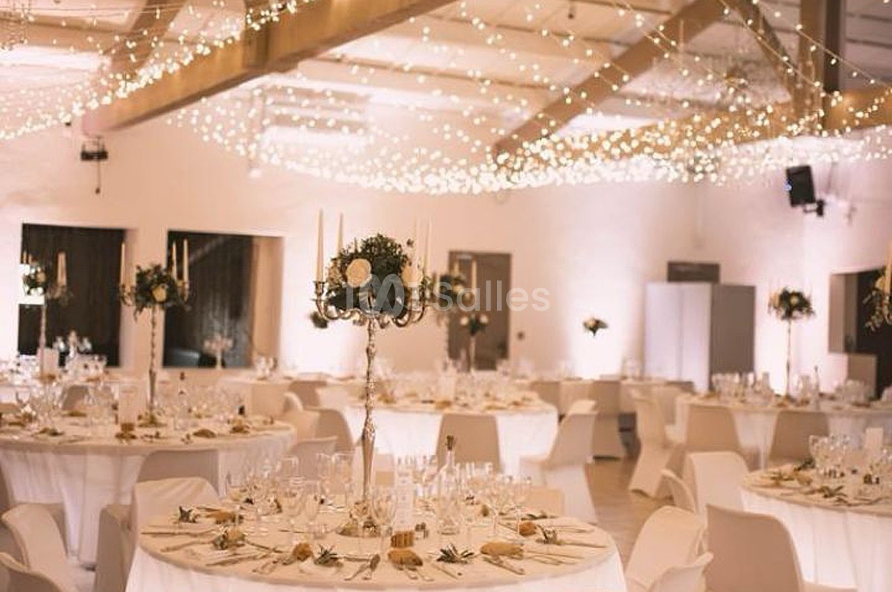 Salle de réception décorée pour un événement, avec tables rondes dressées, chandeliers et guirlandes lumineuses suspendues.