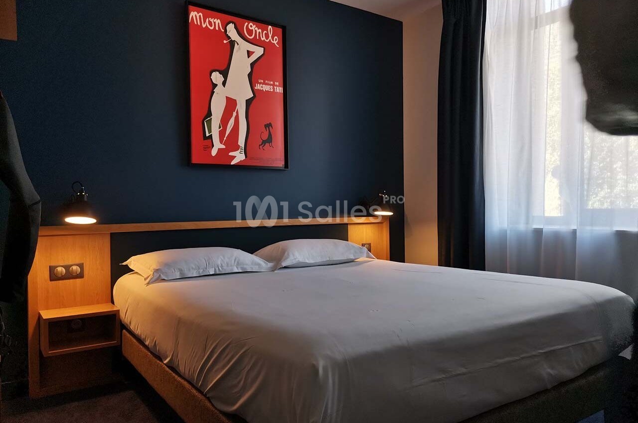Lit double dans une chambre d'hôtel moderne avec mur bleu, éclairage doux et une affiche encadrée au-dessus du lit.