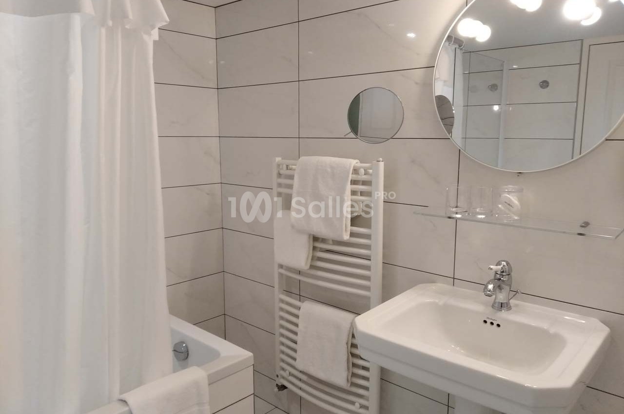 Salle de bain moderne avec carrelage blanc, lavabo, miroir rond, baignoire avec rideau et porte-serviettes chauffant.