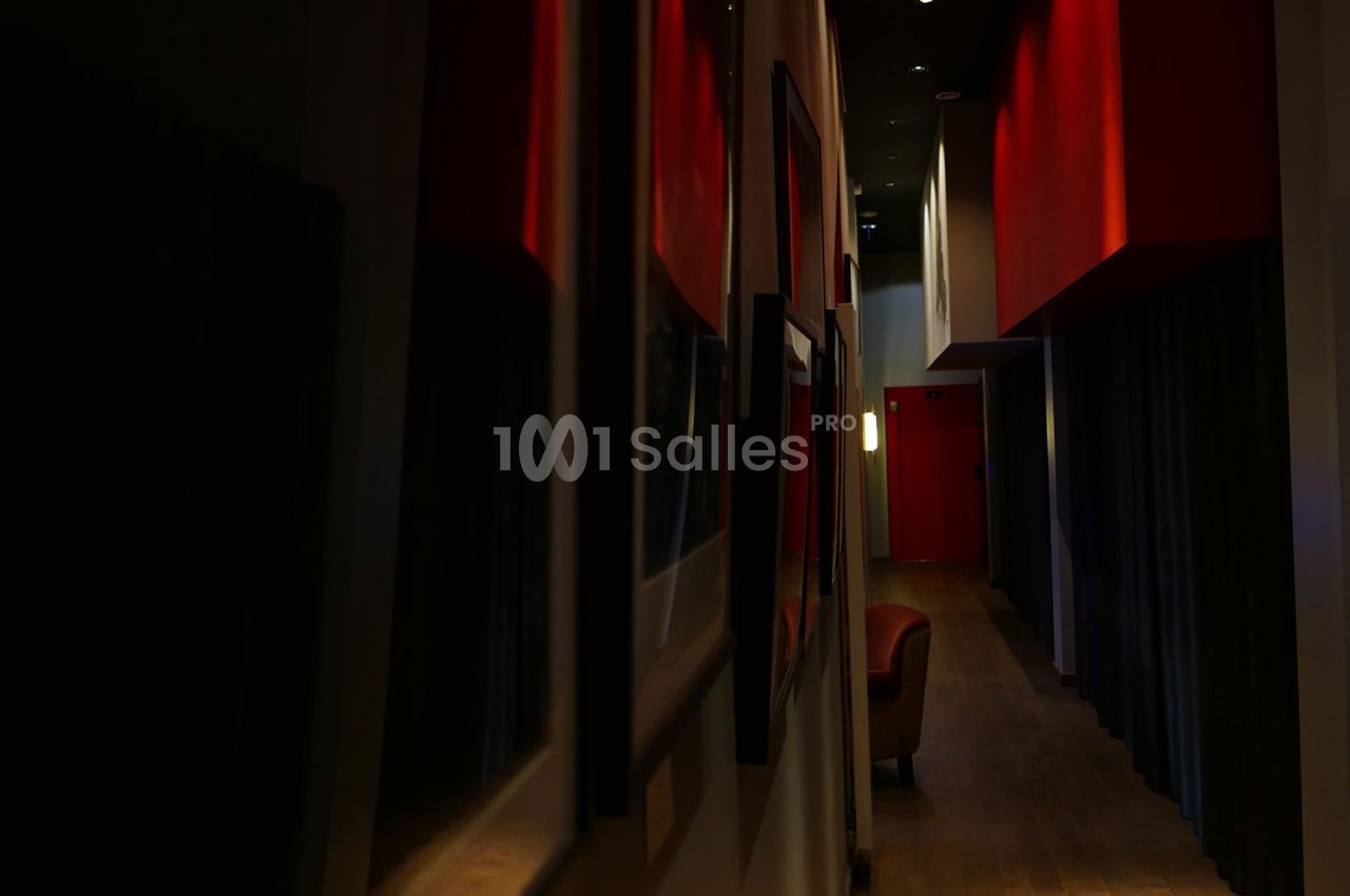 Couloir sombre avec murs rouges, éclairage tamisé et fauteuil visible au fond près d'une porte.