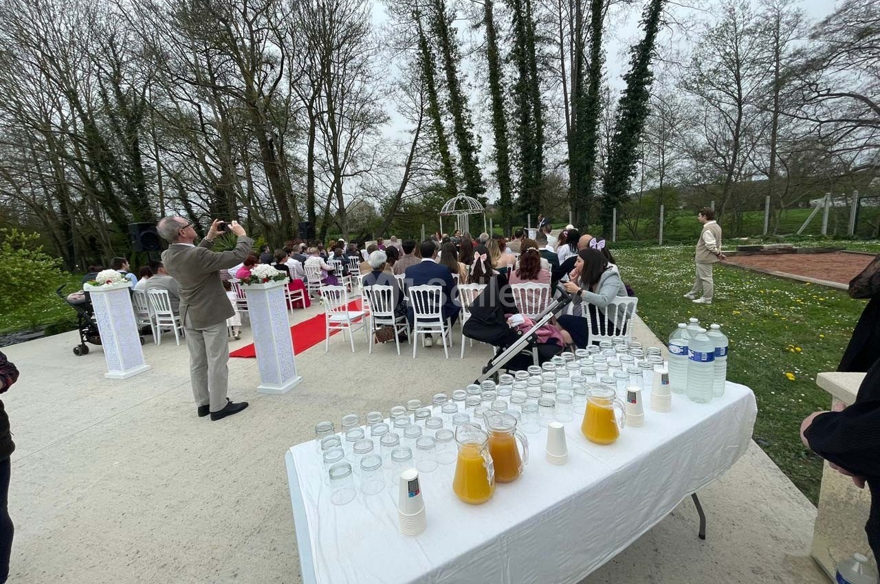 Cérémonie en extérieur avec invités assis, table de boissons au premier plan et arbres en arrière-plan.