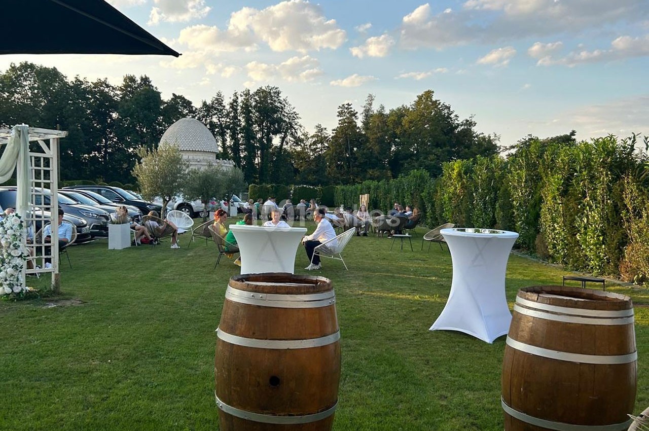 Personnes assises et debout dans un jardin avec des tables hautes, des tonneaux et une coupole blanche en arrière-plan.