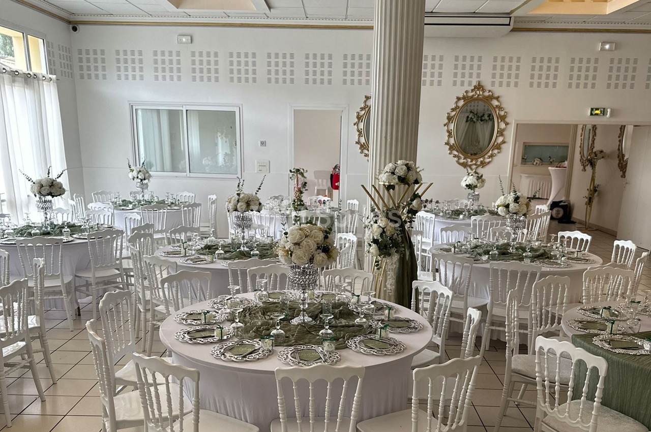 Salle de réception élégante avec tables rondes dressées, décorations florales blanches et chaises blanches.
