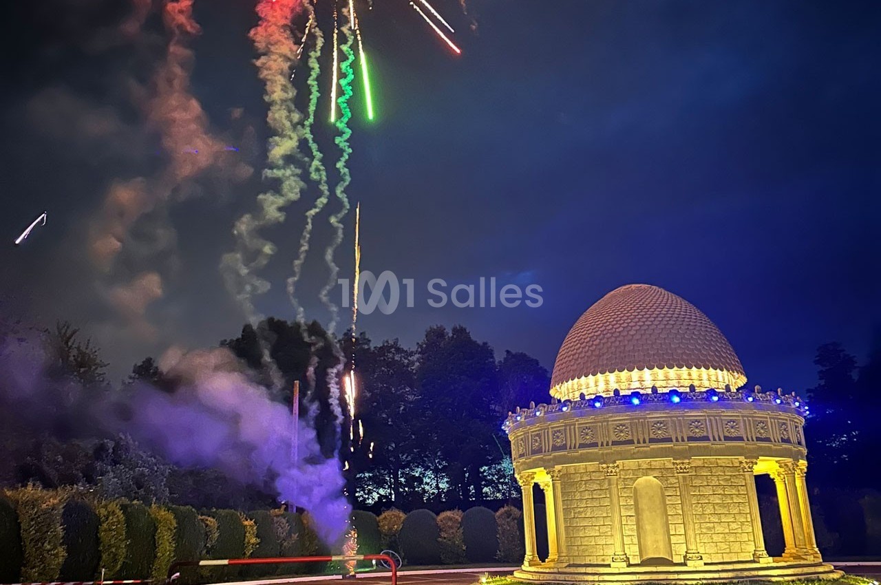 Feu d'artifice illuminant le ciel nocturne au-dessus d'un bâtiment rond éclairé, entouré de végétation.