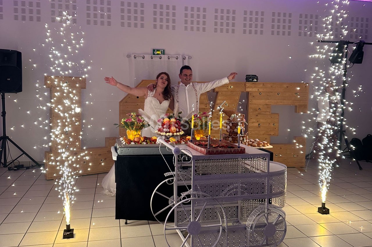 Un couple de mariés souriant pose devant une table décorée de desserts, entourée de fontaines lumineuses.