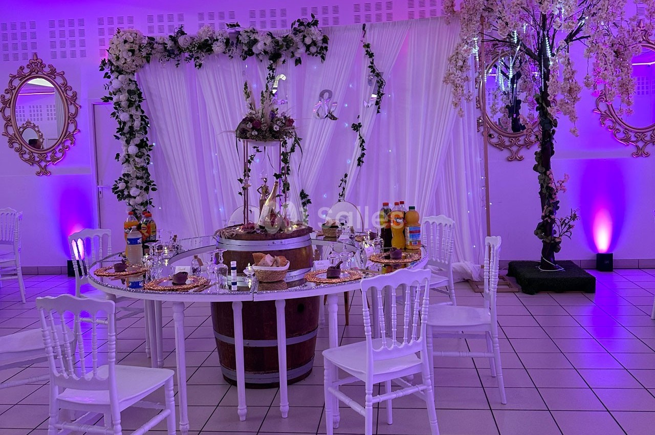 Table décorée pour un événement, entourée de chaises blanches, avec boissons, nourriture et décor floral en arrière-plan.