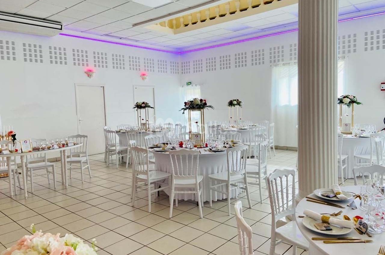 Salle de réception lumineuse avec tables rondes décorées de fleurs, chaises blanches et éclairage LED violet.