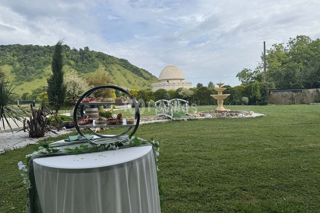Jardin verdoyant avec une table décorée au premier plan, une fontaine et un bâtiment rond en arrière-plan.