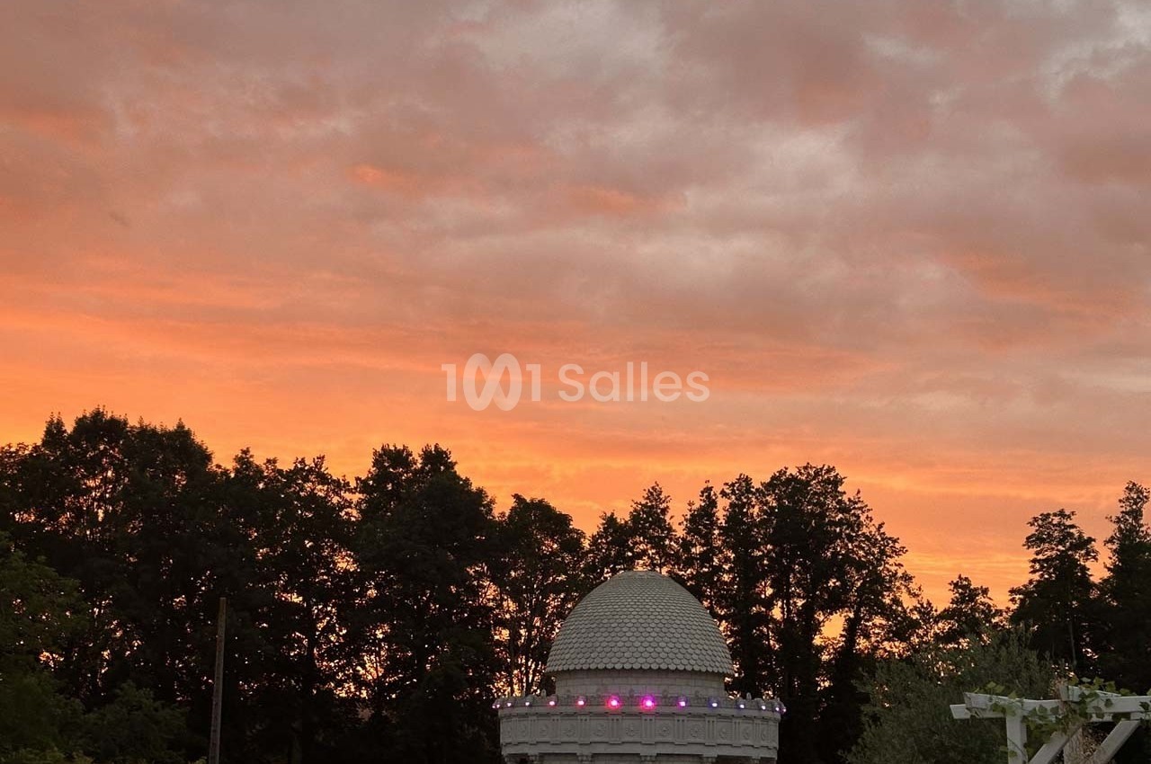 Coupole blanche éclairée au crépuscule, entourée d'arbres sombres sous un ciel orangé nuageux.