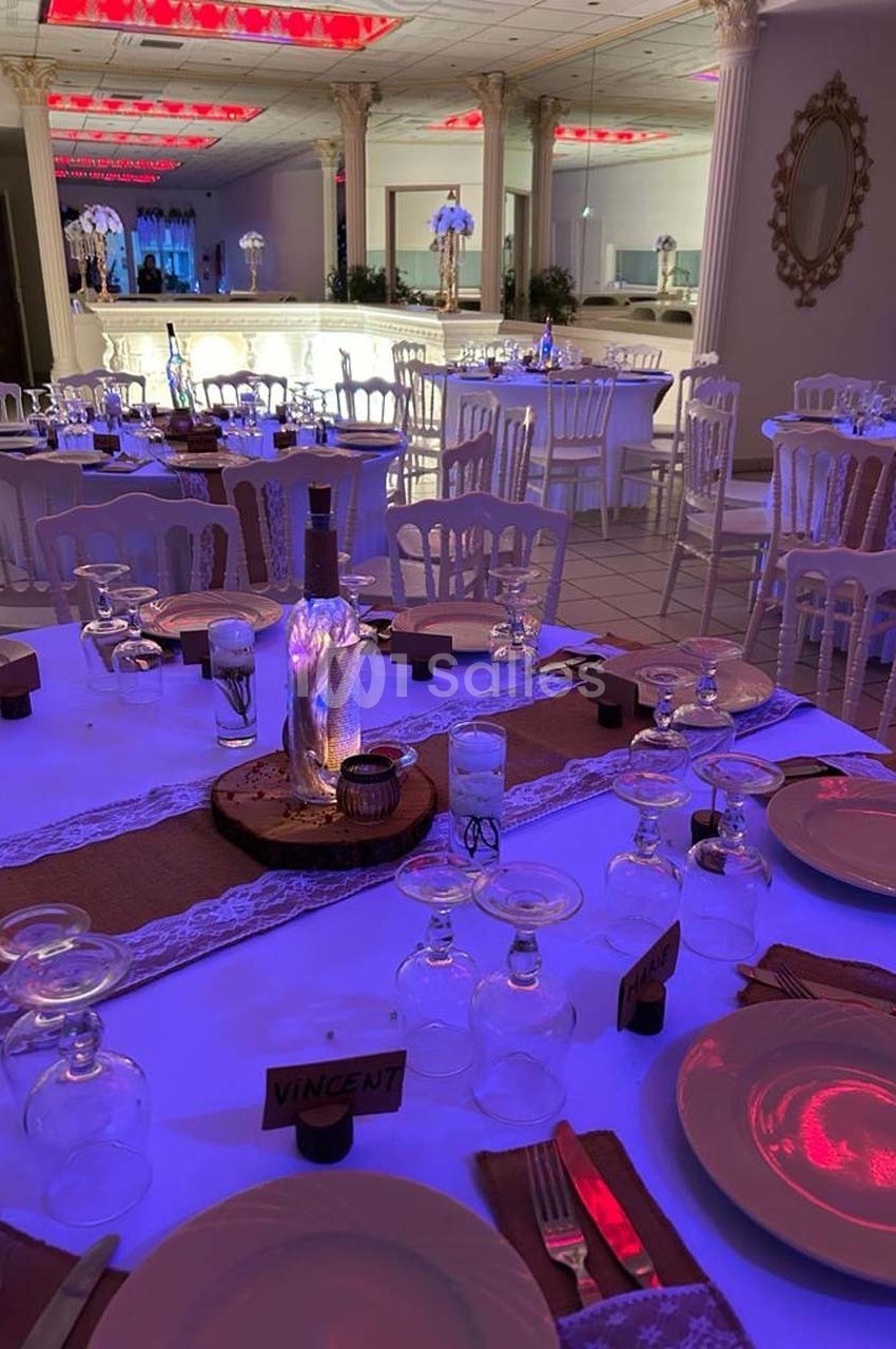Salle de réception décorée avec des tables dressées, nappes blanches, vaisselle et éclairage tamisé aux tons violets.