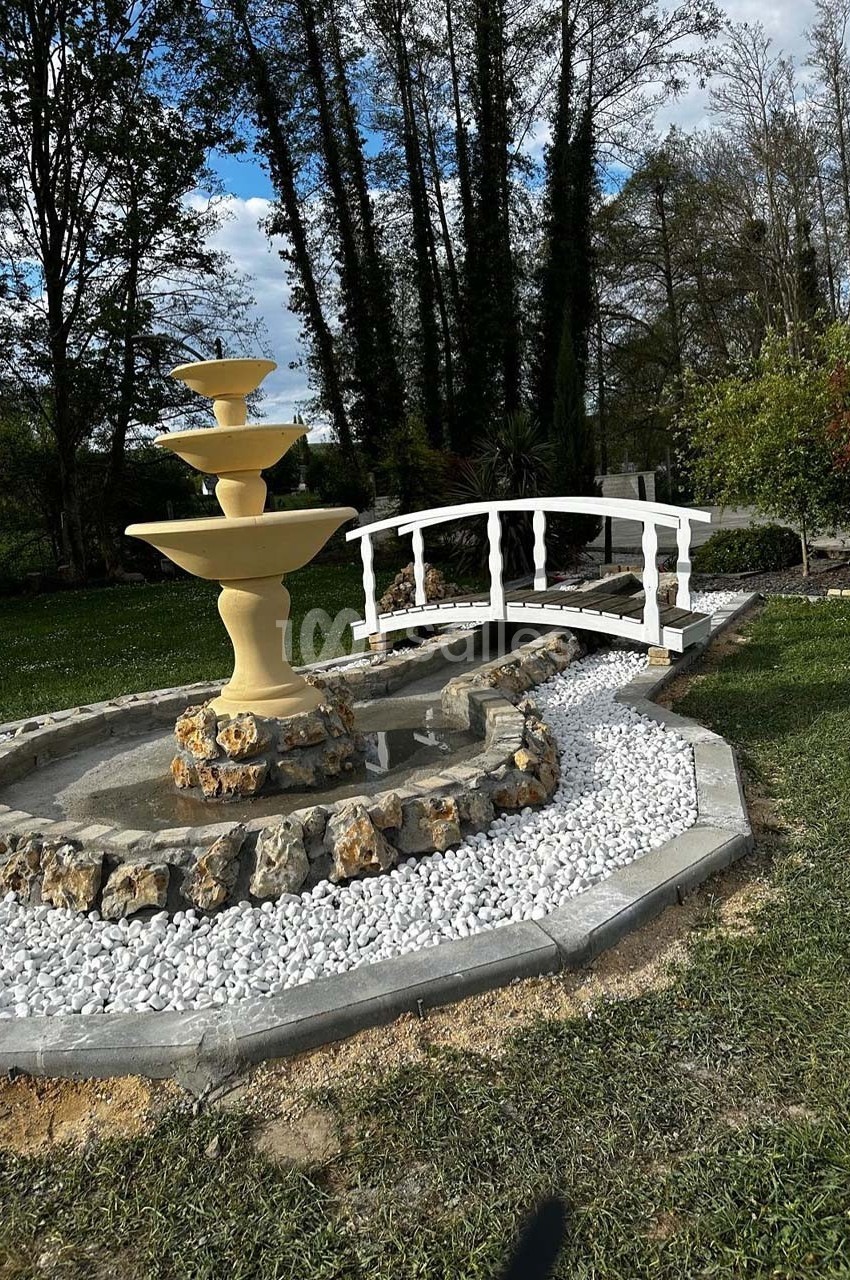 Fontaine beige à trois niveaux entourée de galets blancs, avec un petit pont en bois blanc dans un jardin verdoyant.