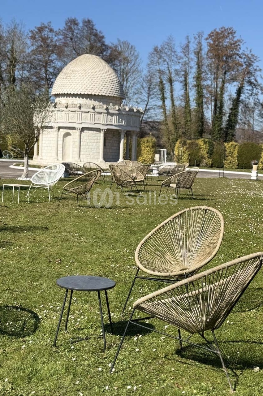 Chaises en rotin et table basse sur une pelouse, avec un pavillon en pierre et des arbres en arrière-plan.