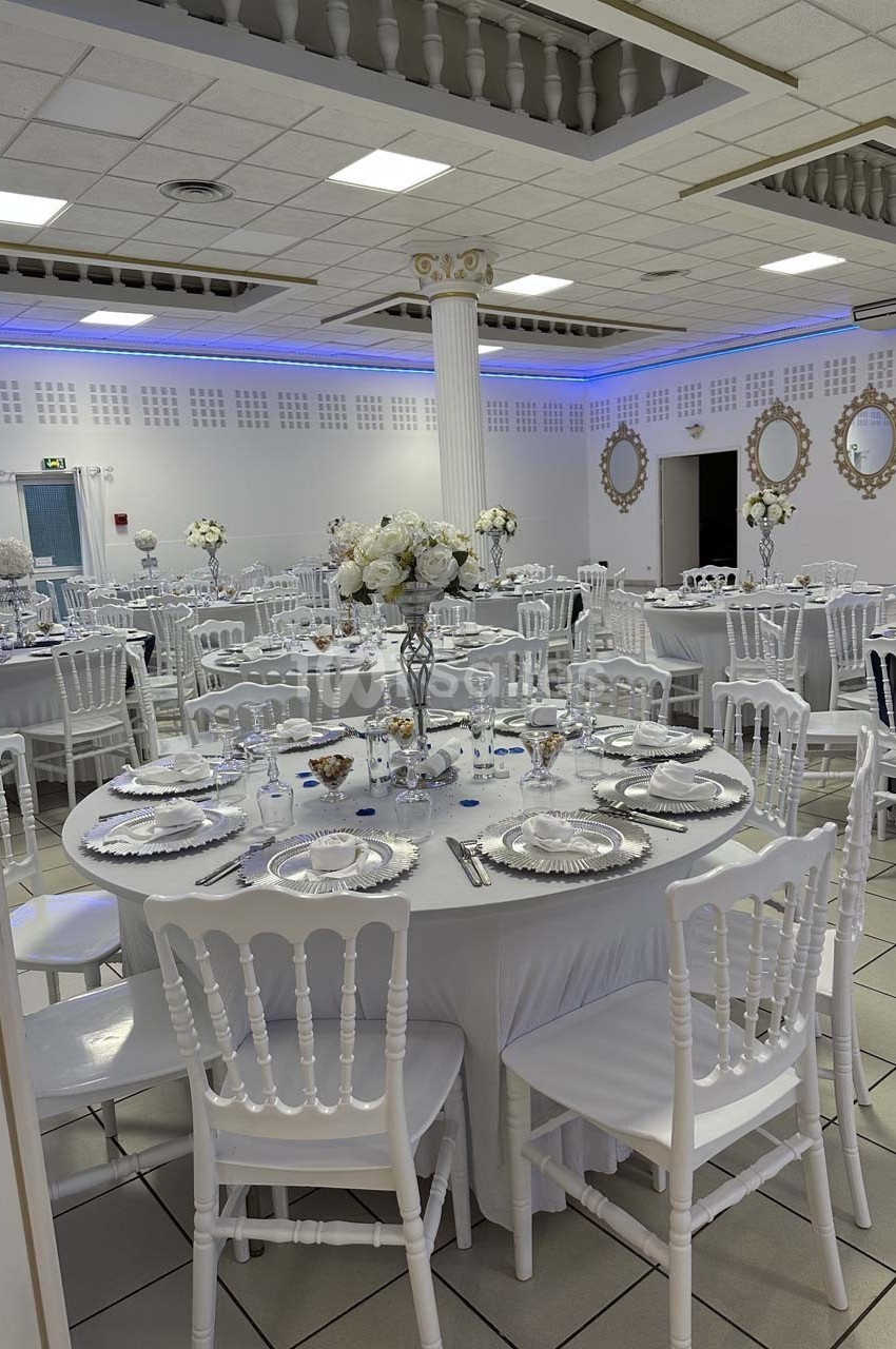 Salle de réception décorée avec des tables rondes dressées, chaises blanches et centre de table fleuris.