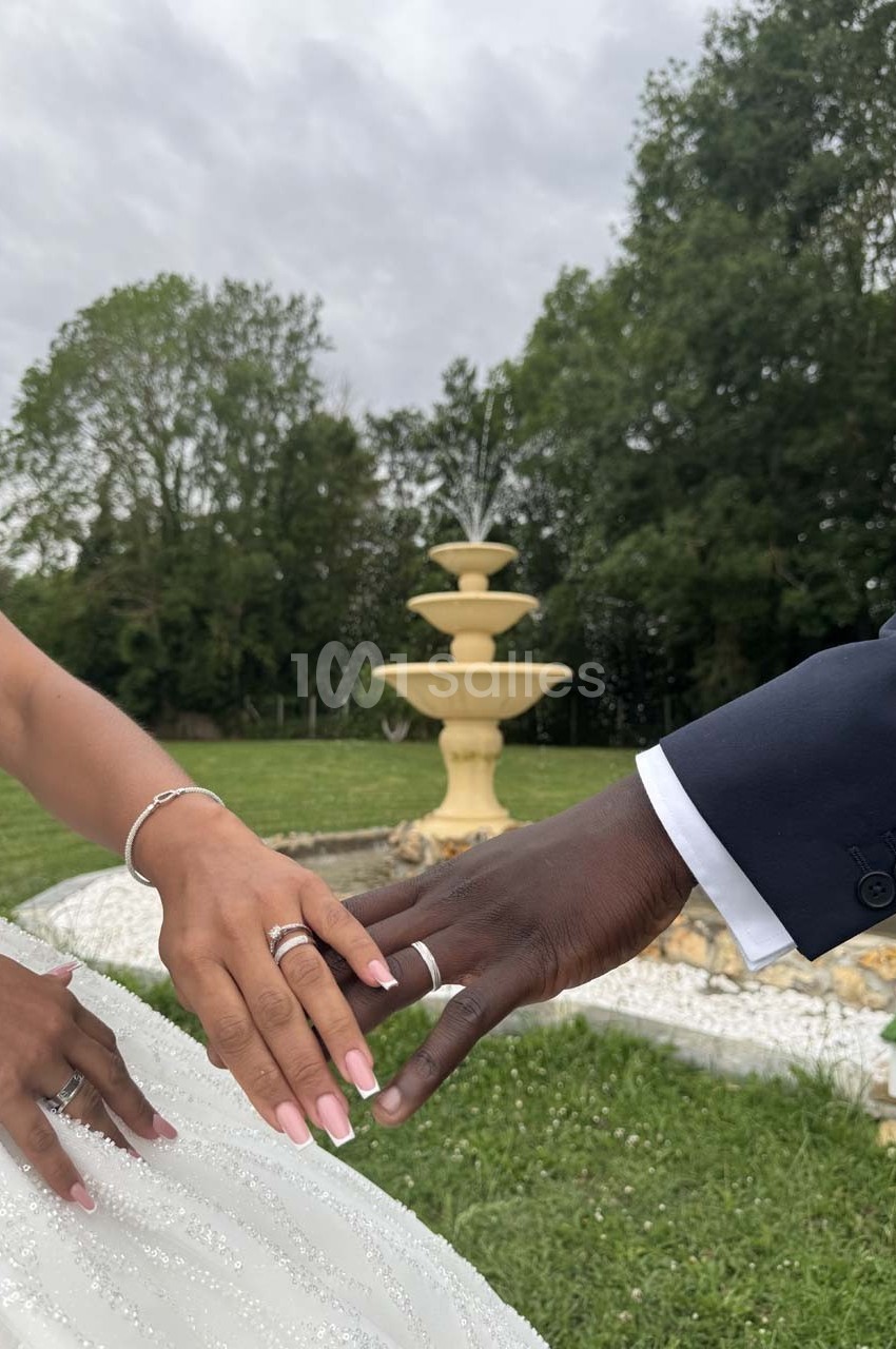 Deux mains, une féminine et une masculine, échangeant des alliances devant une fontaine dans un jardin verdoyant.
