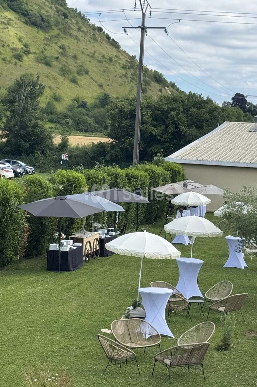Espace extérieur aménagé avec chaises, tables hautes et parasols, entouré de verdure et d'une colline en arrière-plan.