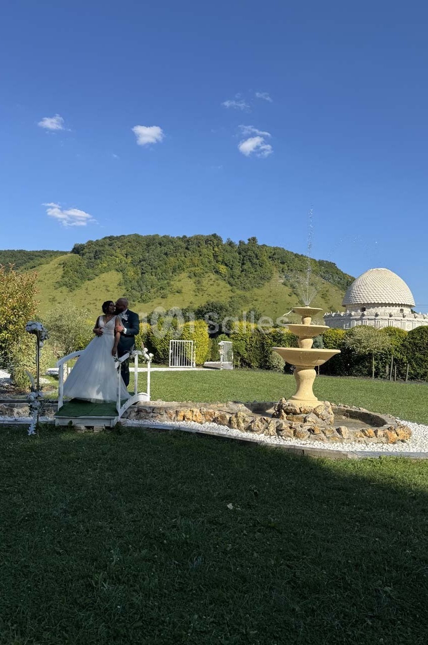 Un couple en tenue de mariage marche dans un jardin verdoyant avec une fontaine et une colline en arrière-plan.
