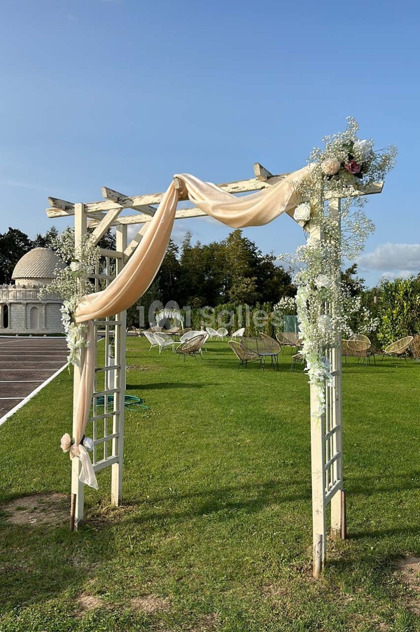 Arche en bois décorée de fleurs blanches et de tissu beige, installée sur une pelouse sous un ciel dégagé.