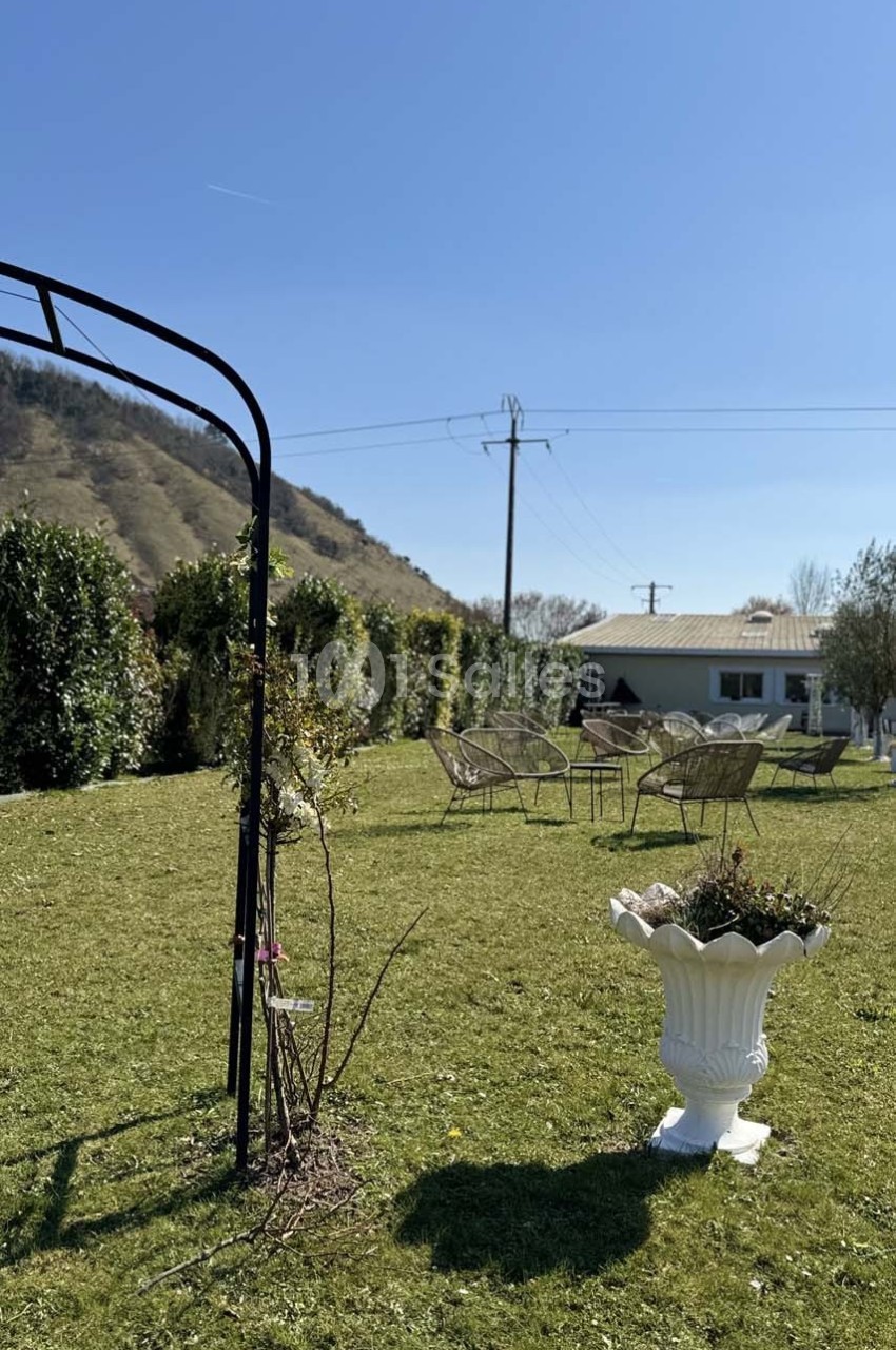 Jardin avec pelouse, pergola métallique, chaises en métal, grand pot blanc et haies devant une maison et des collines.