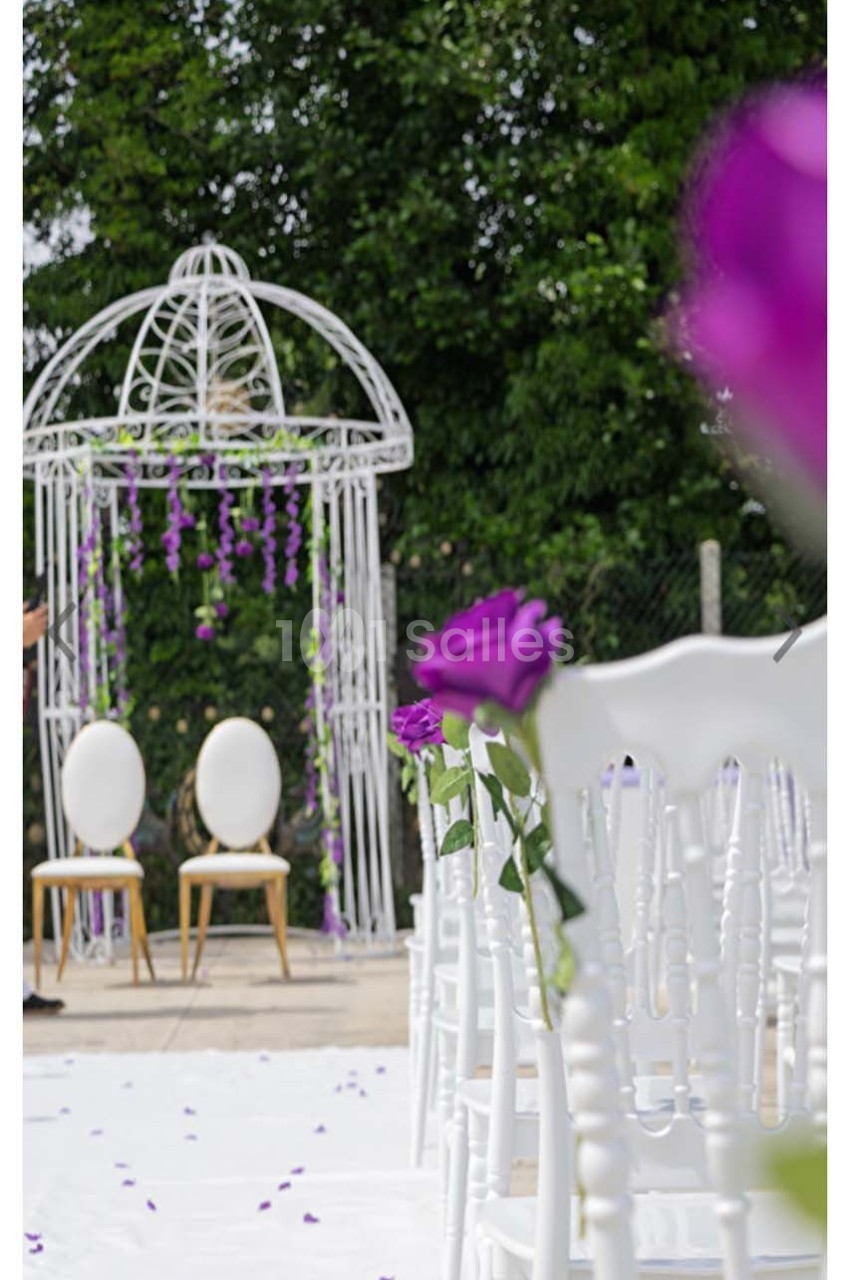 Chaises blanches alignées devant une arche de mariage décorée de fleurs violettes, en extérieur avec arbres en arrière-plan.
