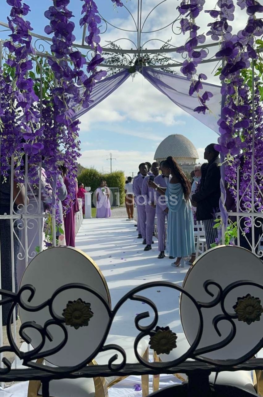 Allée décorée de fleurs violettes menant à une cérémonie en extérieur sous une arche, avec des invités en tenue élégante.