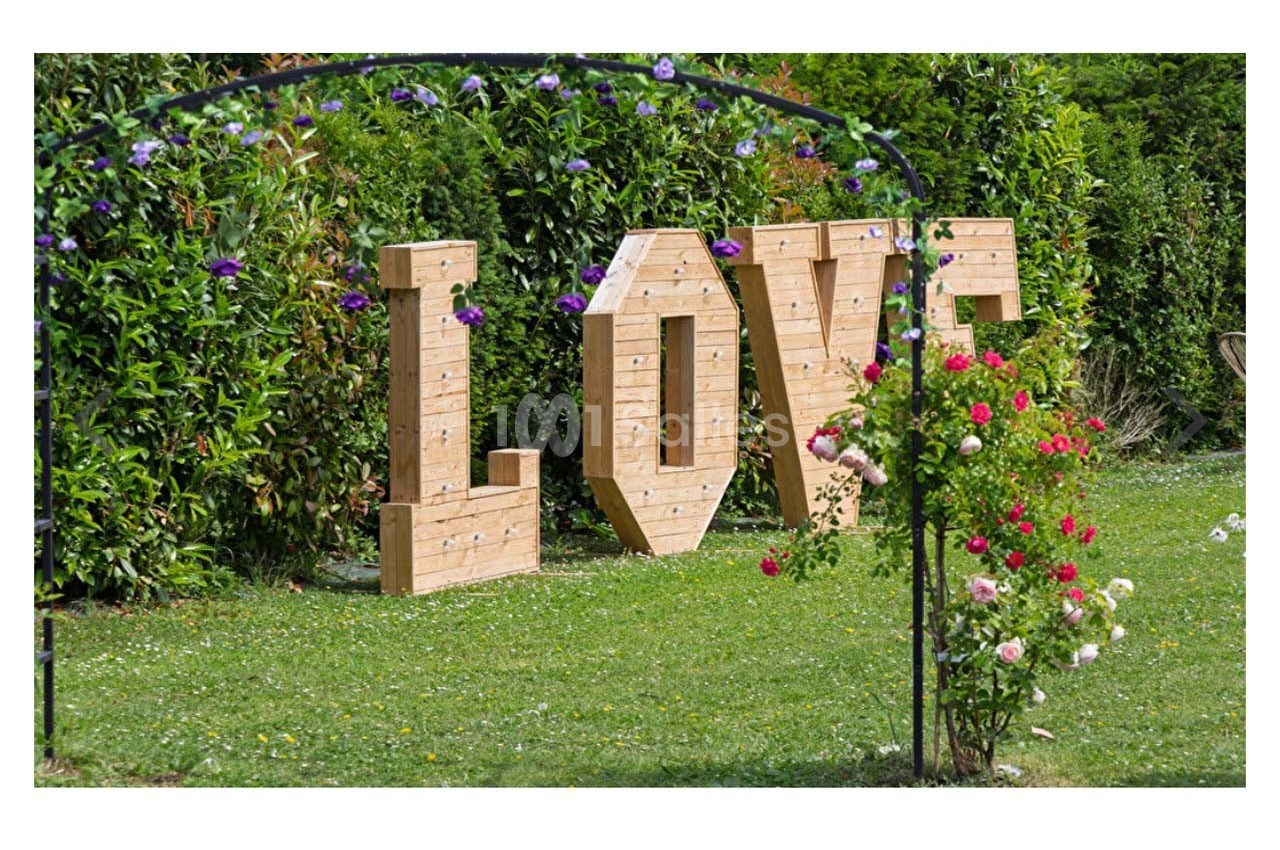 Lettres en bois formant le mot ’LOVE’ dans un jardin verdoyant, entourées de fleurs et d'une arche métallique.