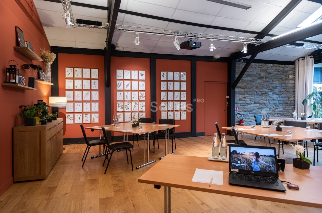 Salle de réunion lumineuse avec tables en bois, chaises noires, mur décoré de cadres et équipements de présentation.